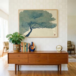 Op de afbeelding: Een rechthoekige ingelijste kunstdruk met een gestileerde blauwgroene boom met donkerblauwe takken tegen een crèmekleurige achtergrond. Het kunstwerk hangt boven een lang houten dressoir met zes lades, een kamerplant, boeken en een kleine ronde klok.