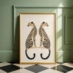 May include: A framed art print featuring two stylized cheetahs facing each other. The cheetahs have tan bodies with black spots and black tails. The artwork is set against a cream background, framed in gold, and leans against a green wall.