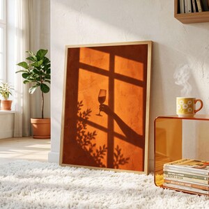 May include: A rectangular framed wall art print featuring a warm orange textured background with the shadow of a window frame, a hand holding a wine glass, and leafy plant silhouettes cast across the surface in a deep brown tone.