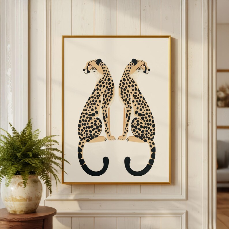 May include: A framed art print featuring two stylized cheetahs facing each other. The cheetahs have tan bodies with black spots and black curved tails. The artwork is set against a cream background and framed in gold. The print is displayed on a cream-colored wall.