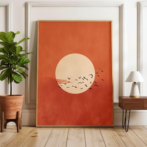 Pode incluir: Uma impressão de arte de parede retangular apresentando um grande círculo de cor creme pálido centralizado contra um fundo laranja queimado sólido e texturizado. Um bando de pequenas silhuetas pretas de pássaros em voo está espalhado pela metade inferior do círculo.