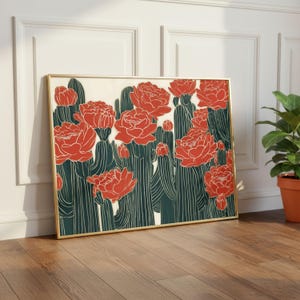 May include: A rectangular wall art print in a thin gold frame featuring a graphic illustration of dark green cacti with prominent vertical lines and large, vibrant orange-red blooming flowers against a light cream-coloured background.
