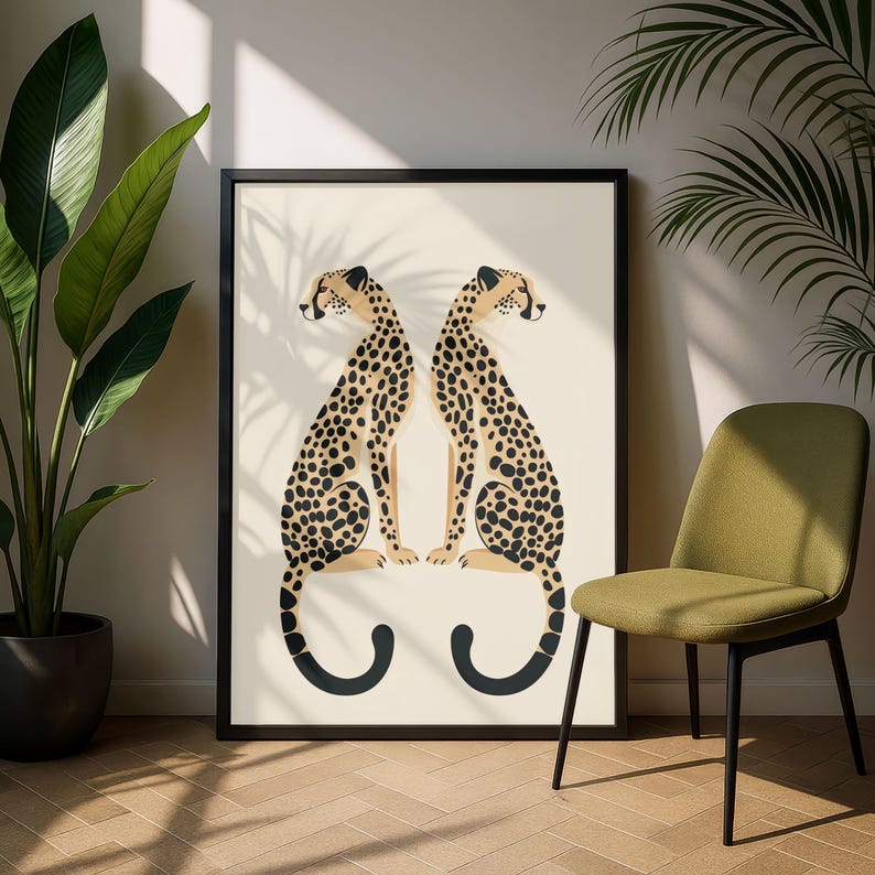 May include: A framed art print featuring two stylized cheetahs facing each other. The cheetahs have tan bodies with black spots and black tails. The artwork is set against a cream background, framed in black, and displayed near a green chair and potted plants.