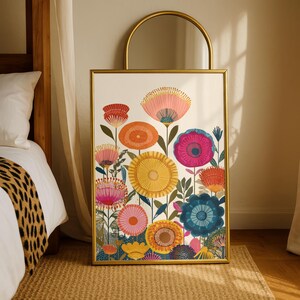 May include: A framed floral art print featuring colorful stylized flowers in shades of pink, orange, yellow, and blue. The artwork is set against a cream background and framed in gold. The print is displayed on a woven rug.