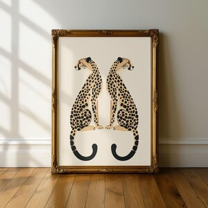 May include: A framed art print featuring two stylized cheetahs facing each other. The cheetahs have tan bodies with black spots and black tails. The frame is gold with ornate detailing. The background is a cream color.