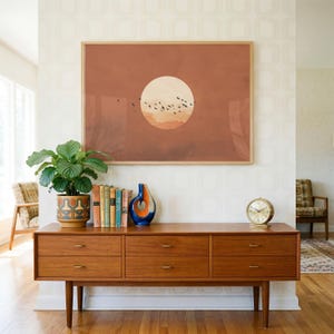 May include: A rectangular wall art print featuring a large, pale circular sun set against a solid terracotta background with a flock of small black birds flying across the centre. The print is framed in a light wood frame and hangs on a wall.