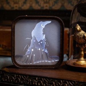 May include: A square, wooden-framed light box with a silhouette of a wizard's hat and a crescent moon. The hat is white and illuminated, with a dark brown frame. The light box sits on a dark wooden surface.