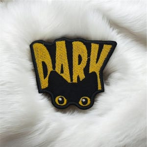 May include: Embroidered patch with the word "DARK" in yellow, stylized lettering. The patch is black with two yellow eye shapes below the text. The patch is on a white, fluffy surface.