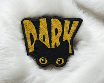 Peut inclure: Écusson brodé noir avec le mot "DARK" en lettres jaunes. Le patch comporte deux yeux jaunes en dessous du texte. L'écusson est posé sur une surface blanche et duveteuse.