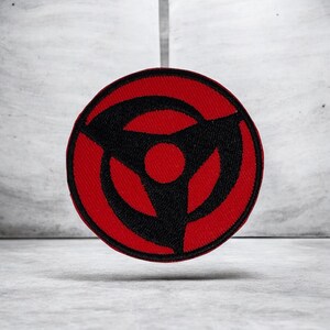 Peut inclure: Un écusson brodé circulaire présentant un fond rouge vif avec un motif noir audacieux en forme de shuriken à trois branches centré autour d'un cercle noir plein. La broderie est dense et uniforme, créant un contraste marqué entre les couleurs rouge et noire sur tout l'emblème circulaire.