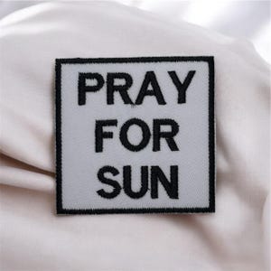 May include: A square patch with the words "PRAY FOR SUN" embroidered in black on a light grey background. The patch has a black border, designed to be sewn or ironed onto clothing or accessories.