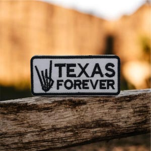 Pode incluir: Um patch retangular branco com uma borda preta e as palavras "TEXAS FOREVER" em letras pretas em negrito. Um gráfico de esqueleto de mão preta está no lado esquerdo do texto. O patch está sobre uma superfície de madeira.