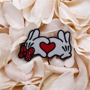 May include: A patch showing two white gloved hands creating a heart shape, with a red heart in the middle. A red bow with white spots is on the left. The patch rests on white flower petals.