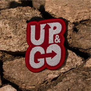 Puede incluir: Un parche rectangular con las palabras "UP & GO" en blanco, delineadas en rojo. El "UP" tiene una flecha hacia arriba, y el "GO" tiene una flecha hacia la derecha. El parche está sobre una superficie de piedra marrón texturizada.