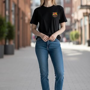 Op de afbeelding: Een zwart t-shirt met korte mouwen en een klein FC Barcelona-embleem op de linkerborst, gecombineerd met een blauwe spijkerbroek en witte sneakers. Het t-shirt heeft een ronde hals en een standaard pasvorm, de spijkerbroek is klassiek blauw.