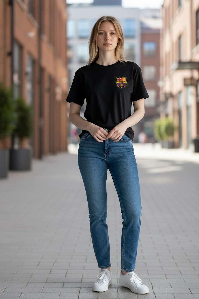 Op de afbeelding: Een zwart t-shirt met korte mouwen en een klein FC Barcelona-embleem op de linkerborst, gecombineerd met een blauwe spijkerbroek en witte sneakers. Het t-shirt heeft een ronde hals en een standaard pasvorm, de spijkerbroek is klassiek blauw.