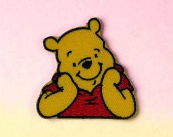 Peut inclure: Un écusson en tissu brodé représentant un ours jaune avec un contour noir, vêtu d'un haut rouge. L'ours est représenté du buste vers le haut avec les deux mains près du visage, mettant en valeur des coutures détaillées et des couleurs vives.
