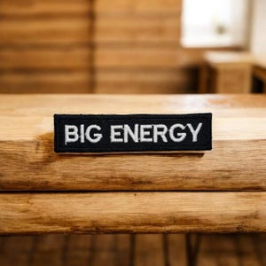 May include: A black rectangular patch with the words "BIG ENERGY" embroidered in white. The patch is placed on a wooden surface with a natural wood grain.