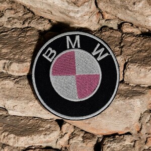 以下が含まれることがあります： BMWのロゴが刺繍された円形のワッペン。黒い外輪、白い文字、そしてマルーンとシルバーグレーのセクションが交互に配置された中央の四分円デザインが特徴で、シルバーグレーのステッチで縁取りされています。