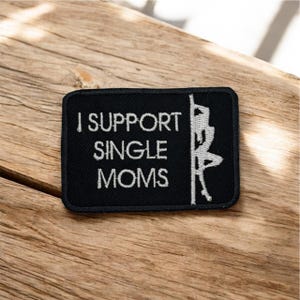 May include: Black rectangular patch with white embroidered text reading "I SUPPORT SINGLE MOMS." A white graphic of a person pole dancing is on the right. The patch is on a wooden surface.