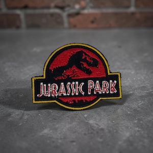 May include: A circular embroidered patch featuring the Jurassic Park logo. The design includes a black silhouette of a Tyrannosaurus Rex skeleton against a red background, with the text JURASSIC PARK in white letters on a black horizontal bar.