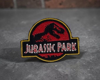 Peut inclure: Un écusson brodé circulaire représentant le logo de Jurassic Park. Le motif comprend une silhouette noire d'un squelette de Tyrannosaurus Rex sur fond rouge, avec le texte JURASSIC PARK en lettres blanches sur une bande horizontale noire.