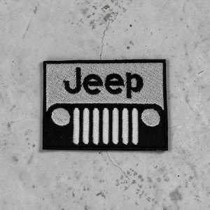 以下が含まれることがあります： ライトグレーの背景に黒い文字でJeepと書かれた長方形の刺繍ワッペン。テキストの下には、7つの垂直スロットと2つの丸いヘッドライトを備えた車両のグリルを黒いグラフィックで表現しています。