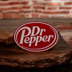 Op de afbeelding: Een rood en wit Dr Pepper-patch. De patch is ovaal van vorm met een rode achtergrond en een witte rand. De woorden "Dr Pepper" zijn in het wit geborduurd, met een klassiek lettertype.