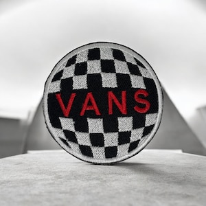 May include: A circular embroidered patch featuring a black and white checkered pattern across the entire surface. The word VANS is stitched in bold red capital letters across the centre of the patch, which has a thick black stitched border.