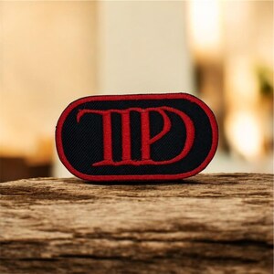 May include: Black and red embroidered patch with the letters "TP" in a stylized font. The patch has a black background with a red border and red lettering. The patch is oval-shaped.