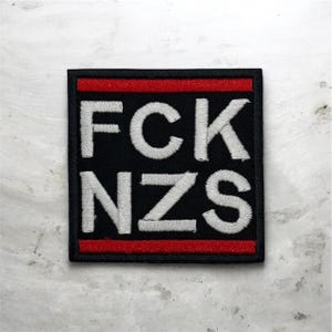 May include: Square black patch with white embroidered text reading "FCK NZS". The patch has a red stripe at the top and bottom, and a black border. The patch is on a light gray surface.
