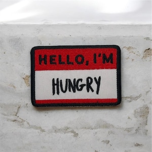 Può includere: Toppa rettangolare con sezione superiore rossa e sezione inferiore bianca. Il testo "HELLO, I'M" è in bianco sulla sezione rossa, e "HUNGRY" è in nero sulla sezione bianca. Bordo nero. Accessorio divertente.
