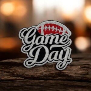 Op de afbeelding: Geborduurde patch met de tekst "Game Day" in witte letters op een zwarte achtergrond. Bovenop staat een rood-witte voetbal. De patch ligt op een houten oppervlak.