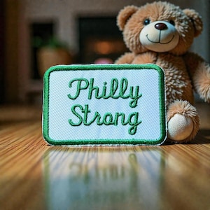 Puede incluir: Un oso de peluche marrón está sentado detrás de un parche blanco con ribete verde. El parche dice "Philly Strong" en letras verdes. El parche está sobre una superficie de madera.