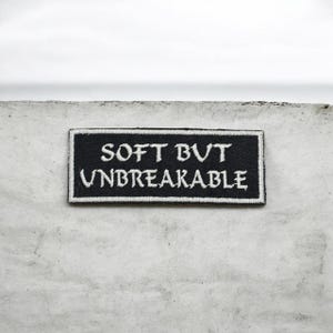 Könnte beinhalten: Ein schwarzer rechteckiger Aufnäher mit weiß gesticktem Text, der „SOFT BUT UNBREAKABLE“ lautet. Der Aufnäher befindet sich vor einem strukturierten, hellgrauen Hintergrund. Der Aufnäher hat einen weißen Rand.