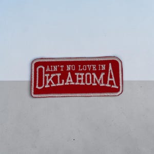 May include: A rectangular embroidered patch with a red background and a white border. The text "AIN'T NO LOVE IN" is stitched in white above the word "OKLAHOMA," which is also stitched in white with larger letters for the O and A.