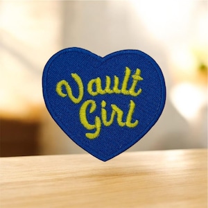 May include: A blue heart-shaped embroidered patch with the words "Vault Girl" in yellow script. The patch is on a light-coloured wooden surface. The patch is a decorative item.
