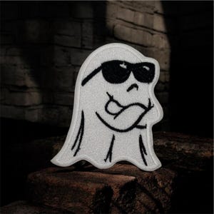 May include: White embroidered patch of a cartoon ghost wearing black sunglasses and with arms crossed. The ghost has a smiling expression. The patch is on a dark, textured surface.