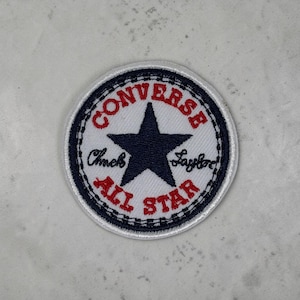 Peut inclure: Un écusson brodé circulaire avec un fond blanc et une étoile bleu foncé au centre. Le texte CONVERSE est en rouge en haut, et ALL STAR est en rouge en bas. Les mots Chuck Taylor sont inscrits en écriture noire au milieu de l'écusson.