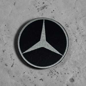 Puede incluir: Un parche bordado circular con fondo negro y un logotipo de estrella de tres puntas de color plateado centrado en el diseño. El parche tiene un borde plateado grueso y cosido que enmarca la forma circular del emblema.