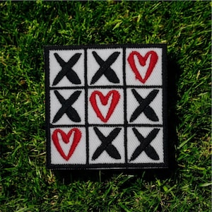 May include: Square patch featuring a tic-tac-toe game design. The grid has black X's and red hearts on a white background, framed by a black border. The patch is likely for clothing or accessories.
