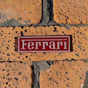 Könnte beinhalten: Ein rechteckiger gestickter Aufnäher mit tiefrotem Hintergrund, einem silberfarbenen Rand und dem Wort Ferrari, das in einer passenden silberfarbenen Schriftart in einer klassischen Serifenschrift in der Mitte aufgestickt ist.