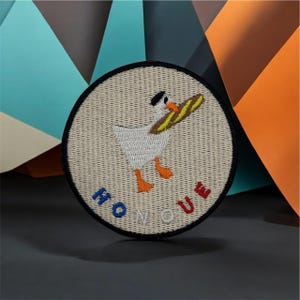 May include: Circular embroidered patch with a cartoon goose wearing a hat and carrying a baguette. The patch reads "NO QUE" in blue and red. It has a black border and a beige background.
