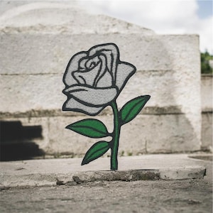 May include: Embroidered patch of a white rose with a black outline, green stem, and green leaves. The patch is set against a blurred, textured background, suggesting an outdoor setting. The rose is the focal point.
