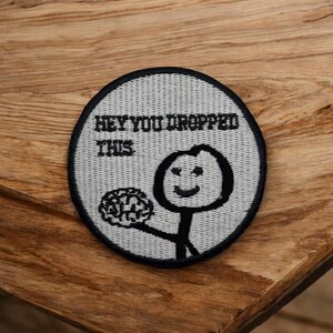May include: A circular patch with a grey background and a black border. It features a stick figure holding a brain and the text "HEY YOU DROPPED THIS."