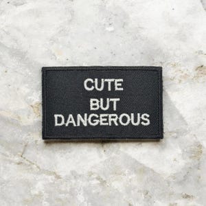 Op de afbeelding: Een zwart rechthoekig embleem met witte geborduurde tekst die luidt: "CUTE BUT DANGEROUS". De patch is waarschijnlijk van stof en heeft een gestikte rand. De achtergrond is een lichtgrijs, getextureerd oppervlak.