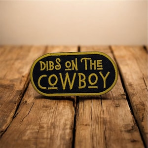 Pode incluir: Patch bordado preto e dourado com o texto "Dibs on the Cowboy". O patch tem um fundo preto com letras douradas e uma borda dourada. O patch tem formato oval e está sobre uma superfície de madeira.