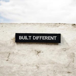 Pode incluir: Um patch retangular preto com as palavras "BUILT DIFFERENT" bordadas em branco. O patch está colocado sobre uma superfície texturizada esbranquiçada, com um céu nublado ao fundo. O patch é uma declaração simples e ousada.