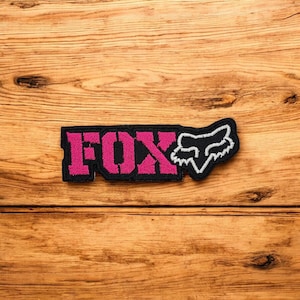 Puede incluir: Parche bordado negro y rosa con la palabra "FOX" en letras rosas y el logotipo de Fox Racing en negro y blanco. El parche está sobre un fondo de madera.