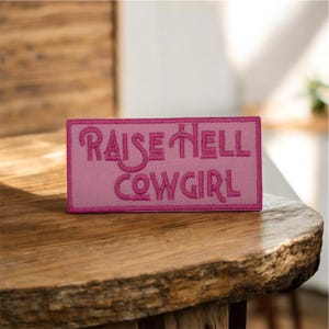 May include: A rectangular pink patch with the embroidered phrase "RAISE HELL COWGIRL" in a darker pink. The patch is likely made of fabric and features a detailed, stylized font. The patch is displayed on a wooden surface.